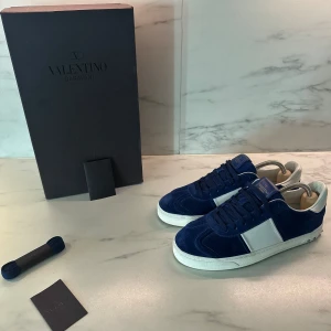 Blå och vita sneakers från Valentino - Snygga blå sneakers från Valentino med vita detaljer. Skorna har en klassisk design med snörning och är tillverkade i mocka. Perfekta för en stilren look. Alla nitar kvar, strl 41.5 skick 9/10 Nypris runt 8000 Box, kort & snören medföljer