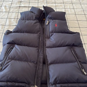 Svart dunväst från Ralph Lauren - Säljer en snygg svart barn dunväst från Ralph Lauren med dragkedja och två fickor framtill. Perfekt för kyligare dagar och ger en stilren look. Västen har en diskret logga på bröstet.