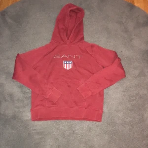 Röd hoodie från GANT - Säljer en snygg röd hoodie från GANT med ett tryck av deras logga och ett sköldemblem på bröstet. Tröjan har en klassisk huva och långa ärmar, perfekt för en avslappnad stil. Passar bra till både jeans och joggers.