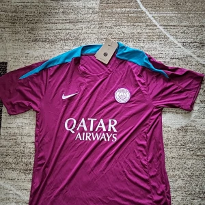 Fotbollströja från PSG - Säljer en snygg lila fotbollströja från Nike med turkosa detaljer på axlarna. Tröjan har korta ärmar och ett tryck av Qatar Airways på framsidan. Perfekt för fotbollsfans som vill ha något unikt i garderoben.