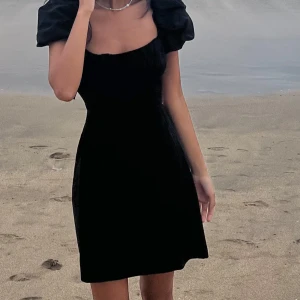 Svart klänning med puffärmar - Snygg svart klänning med puffärmar och öppen rygg. Den har en elegant snörning baktill som ger en unik touch. Perfekt för en stilren look vid stranden eller en kväll ute.