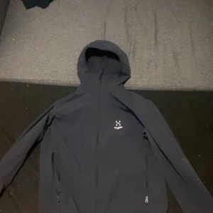 Svart jacka med huva från Haglöfs - Säljer en stilren svart jacka från Haglöfs med huva och dragkedja framtill. Perfekt för utomhusaktiviteter med sin funktionella design. Jackan har långa ärmar och en diskret logga på bröstet.