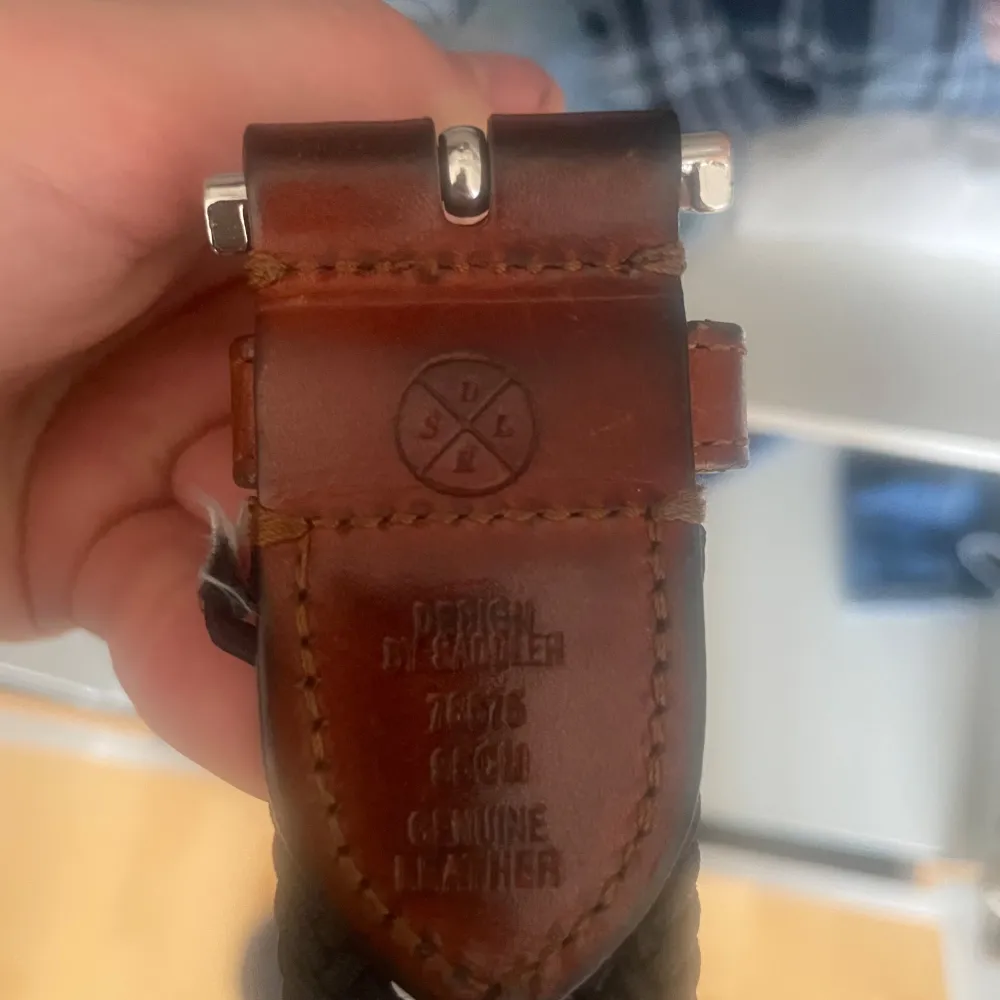Snyggt blått flätat bälte med bruna läderdetaljer och silverfärgat spänne. Perfekt för att ge en stilren touch till din outfit. Bältet är tillverkat av genuint läder och har en klassisk design.. Asusteet.