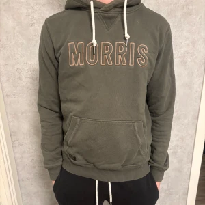 Grön hoodie från Morris - Säljer en snygg grön hoodie från Morris med broderad logga på bröstet. Tröjan har en klassisk känguruficka och justerbar huva med vita snören. Perfekt för en avslappnad stil.