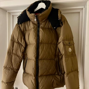 Moncler himalaya jacket - Säljer en snygg brun dunjacka från Moncler. Jackan har en ficka på ärmen med Moncler-loggan. Insidan är i en cool röd färg. Perfekt för kylan.
