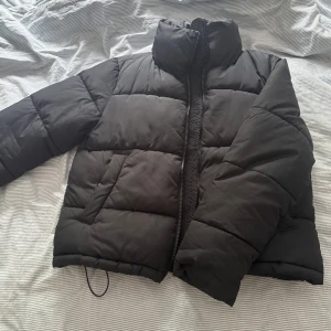 Svart pufferjacka med dragkedja - Säljer en svart pufferjacka med hög krage och dragkedja framtill. Jackan är quiltad och har en loose passform, perfekt för kalla dagar. Enkel och stilren design som funkar till det mesta. Snygg och bekväm att bära under höst och vinter.