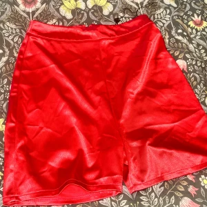 Röd satin-kjol från Club L London - Snygg röd kortkjol i glansigt satinmaterial från Club L London. Kjolen har en liten slits framtill och dragkedja bak för enkel påklädning. Perfekt för dig som vill sticka ut med en färgstark look.