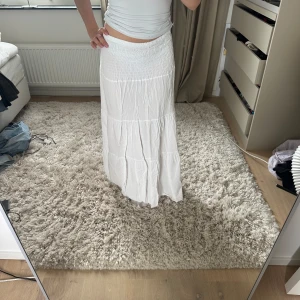 Vit långkjol med smock i midjan - Säljer en superfin vit långkjol med smockad midja och volangiga partier. Kjolen är luftig och har en flowy känsla, perfekt för varma dagar. Enkel att matcha med crop tops eller linnen för en chill och fräsch look.