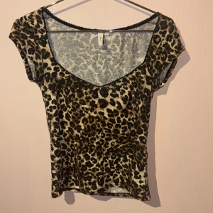 Leopardmönstrad topp från Nelly XS - Snygg topp från Nelly i leopardmönster med v-ringning och svarta spetsdetaljer längs halsringningen. Toppens färger går i beige, brunt och svart och den har korta ärmar. Perfekt för dig som vill sticka ut med ett djurmönstrat plagg.