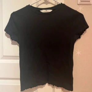 Svart ribbad topp från H&M - Svart ribbad topp från H&M med korta ärmar och rund hals. Toppen har vågiga kanter vid ärmslut och nederkant som ger en extra touch. Materialet är mjukt och stretchigt, perfekt för att matcha med jeans eller kjol. Enkel och stilren design.