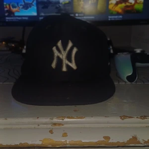 Svart New York Yankees keps med blommor - Snygg svart keps från New York Yankees med broderade blå blommor runt det klassiska NY-loggan framtill. Platt skärm och cool streetstil, perfekt för att lyfta din outfit med en unik detalj. Strl 58.7är prutbar vid snabbaffär 
