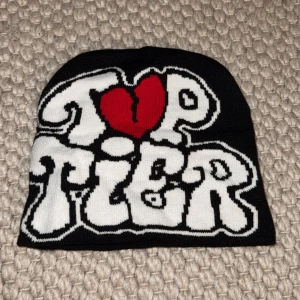 Svart mössa med Top Tier-tryck - Cool svart mössa med vit text 'Top Tier' i stor, bubblig stil och ett rött hjärta som O. Perfekt accessoar för att sticka ut i mängden. Mjuk och stretchig, passar de flesta huvudstorlekar.