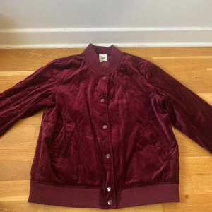 Vinröd sammet bomberjacka från Vans - Säljer en vinröd bomberjacka från Vans i mjuk sammet. Jackan har klassisk bomberkrage, knappar framtill och två fickor. Broderad Vans-logga på bröstet . Perfekt för dig som vill sticka ut med en snygg färg och coolt material.