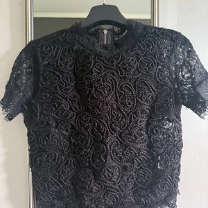 Svart spetsblus med dragkedja - Unik svart blus med korta ärmar i spets och broderade detaljer framtill. Baksidan har en snygg dragkedja som ger en cool touch. Perfekt för dig som vill sticka ut med en elegant men edgy look.