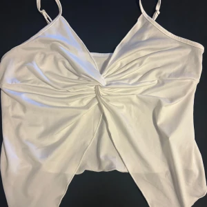 Vit omlott linnetopp med knut - Snygg vit linnetopp med smala axelband och en cool knutdetalj framtill som ger en omlott-effekt. Toppen är croppad och har en lätt glansig finish, perfekt för att lyfta vilken outfit som helst. Materialet känns mjukt och följsamt mot huden. Öppen mage strl xl men funkar l