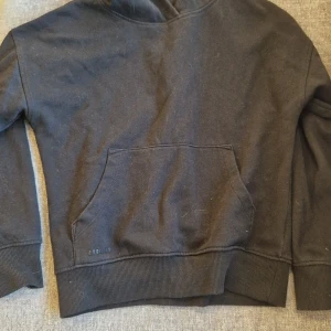 Svart hoodie från Wawai med ficka - Svart hoodie från Wawai i storlek 134/140. Tröjan har en klassisk huval. Tillverkad i mjuk bomull med ribbade muddar vid ärmslut och nederkant. Perfekt för chill dagar och enkel att matcha med jeans.