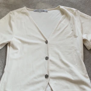 Krämvit v-ringad blus med knappar - Snygg krämvit blus från 157 i storlek S. Den har en djup v-ringning och tre coola mörka knappar framtill. Materialet känns mjukt och lätt, perfekt för dig som gillar enkel och clean stil. Kortärmad och med en rak passform som är lätt att matcha.