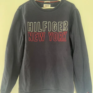 Mörkblå Hilfiger Denim tröja M - Mörkblå sweatshirt från Hilfiger Denim med stor vit och röd text 'HILFIGER NEW YORK' på bröstet. Klassisk rund hals, långa ärmar och broderad logga vid ärmslut. Skön bomullsmix som passar perfekt till jeans eller joggers.