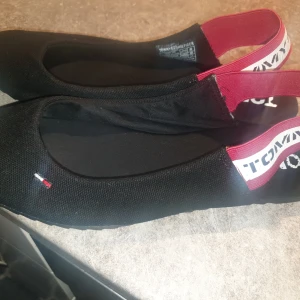 Svarta ballerinaskor Tommy Jeans - Svarta ballerinaskor från Tommy Jeans med elastiskt band i vinrött och vitt med logga. Skorna har rund tå och är tillverkade i textilmaterial. Liten broderad logga framtill ger en clean och sportig look.