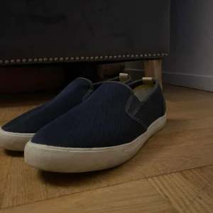 Mörkblå slip-on sneakers från Lyle & Scott - Snygga mörkblå slip-on sneakers från Lyle & Scott med vit sula och gul broderad logga på hälen. Ovandelen är i textil med diskret struktur och insidan har gula detaljer. Perfekta för en clean och avslappnad stil.