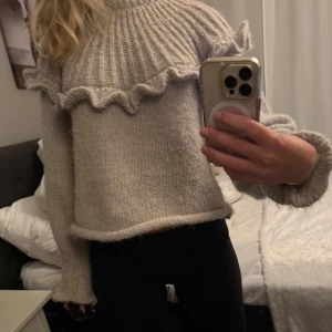 Beige stickad tröja med volang  - Superfin beige stickad tröja med bred volangdetalj runt axlarna och ribbad struktur upptill. Tröjan har lång ärm och en lite loose passform, perfekt för dig som gillar en mysig och trendig look. Passar perfekt till jeans eller kjol för en chill vibe.