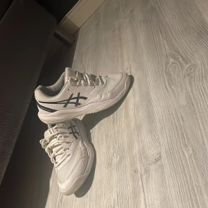 Vita Asics sneakers strl 40,5 - Säljer ett par vita Asics sneakers med svarta detaljer och logga på sidan. Skorna har snörning, rund tå och är tillverkade i syntet och mesh för bra andningsförmåga. Perfekta för dig som gillar sportig stil och vill ha bekväma skor till vardagen.