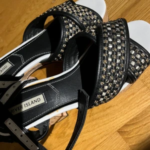 River Island svarta högklackade sandaler med nitar - Snygga svarta sandaler från River Island med breda remmar dekorerade med små nitar i guld och silver. Skorna har öppen tå, vit innersula och justerbart spänne runt ankeln. Perfekta för dig som vill sticka ut med coola detaljer och en trendig look. Står ingen stl men skulle uppskatta att de är stl 38-39.
