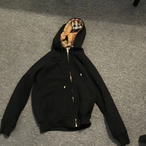 Svart hoodiejacka från Burberry M - Svart hoodiejacka från Burberry med dragkedja och klassiskt rutigt foder i huvan. Jackan har snörning i huvan och ribbade muddar. Perfekt för dig som vill ha en stilren och lyxig look med ikoniska Burberry-detaljer.