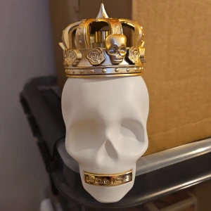 Police To Be The King EdP - Parfymflaska formad som en vit dödskalle med en detaljerad guldfärgad krona på toppen och gulddekor vid munnen där det står 'Police'. Flaskan är tillverkad i matt glas och har en unik, edgy design som sticker ut i hyllan.