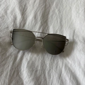 Silvriga solglasögon med metallbåge - Snygga solglasögon med rundade, mörkgrå linser och en unik silvrig metallbåge. Bågen har en cool, geometrisk design med dubbla linjer över glasen och tunna skalmar. Perfekt för dig som gillar en modern och edgy stil.