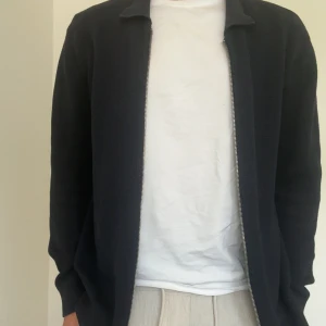 Svart stickad cardigan jacka med dragkedja - Snygg svart stickad cardigan jacka med hel dragkedja framtill och klassisk krage. Jackan har en clean och stilren look, perfekt att slänga på sig över en t-shirt. Långärmad och i mjukt material som känns skönt mot huden.