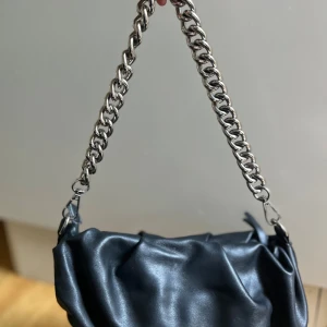 Svart axelväska med kedja - Snygg svart axelväska i mjukt skinnliknande material med cool grov silverfärgad kedja som axelrem. Väskan har en slouchy, puffig form och stängs med dragkedja. Perfekt för dig som gillar en edgy och trendig look.