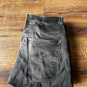 Grå jeans från Nudie Jeans Co - Säljer ett par grå jeans från Nudie Jeans Co med klassisk femficksdesign och snygga detaljer på bakfickan. Jeansen har en smal passform och är tillverkade i mjukt denimtyg. Perfekta för dig som gillar en stilren och avslappnad look.