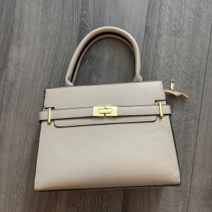 Beige handväska med gulddetaljer - Stilren beige handväska i skinn med guldfärgade metalldetaljer och klassisk låsdetalj framtill. Väskan har både handtag och avtagbar axelrem, samt rymligt fack med dragkedja och innerficka. Perfekt för dig som gillar en clean och elegant look. Väskan är helt oanvänd och ny🥰