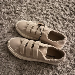 Beige sneakers med fransade kanter - Snygga beige sneakers från Even&Odd med coola fransade kanter och tre breda kardborreband. Skorna har en platt sula och är tillverkade i bomullstyg. De är aldrig använda och har därför prislappen kvar. 