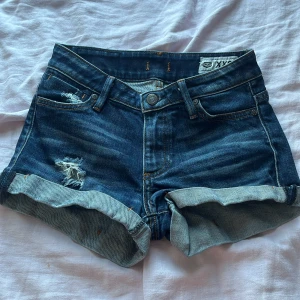 Blå jeansshorts från Crocker XXS - Snygga blå jeansshorts från Crocker i storlek XXS med uppvikta ben och slitna detaljer framtill. Klassisk femficksmodell, knapp och dragkedja i midjan. Perfekt för dig som gillar en chill och trendig look på sommaren.