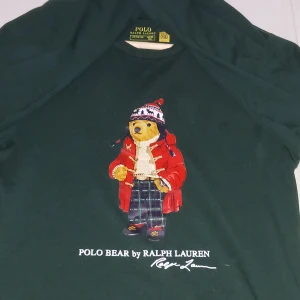 Mörkgrön Polo Bear tröja Ralph Lauren - Säljer en mörkgrön Polo Bear tröja från Ralph Lauren i storlek 14-16 år. Tröjan har en stor tryckt nalle med röd jacka och mönstrad mössa på framsidan. Rund hals och normal passform, tillverkad i mjuk bomull. Perfekt för dig som gillar klassisk streetstyle med en twist.