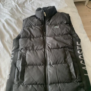 Svart dunväst från Lacoste - Snygg svart dunväst från Lacoste med broderad krokodil-logga på bröstet och stort Lacoste-tryck längs sidan. Västen har hög krage, dragkedja framtill och två fickor. Perfekt att slänga över en hoodie för en sportig look. Passar bra till M.