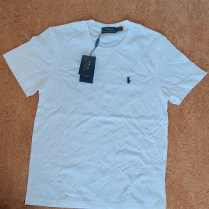 Vit t-shirt från Polo Ralph Lauren - Klassisk vit t-shirt från Polo Ralph Lauren med broderad logga på bröstet. T-shirten har rund halsringning och korta ärmar. Tillverkad i mjuk bomull för en skön och stilren look. Perfekt till jeans eller shorts för en clean vibe.