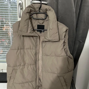 Beige dunväst från 157, XS - Säljer en beige dunväst från 157 i storlek XS. Västen har hög krage, två fickor och stängs med dragkedja. Den är quiltad och har en clean, enkel look som passar perfekt till lager-på-lager-stil under kyliga dagar.