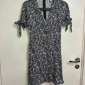 Svartvit blommig kortklänning Amisu - Supersöt kortklänning från Amisu med svart botten och vitt blommönster. Klänningen har v-ringad hals, korta ärmar med knyt och en lätt figurnära passform. 