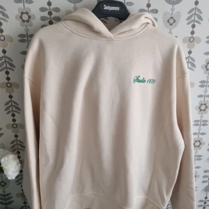 Beige hoodie Studio 1971 - Snygg beige hoodie från Studio 1971 med broderad grön text på bröstet. Mjuk bomullskänsla, huva och klassisk passform. Perfekt för en chill och avslappnad stil.