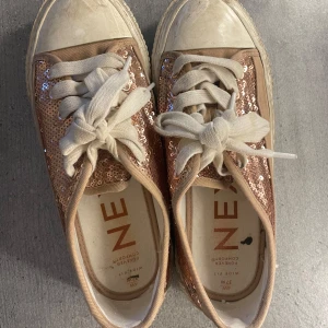 Glittriga sneakers från Next - Snygga sneakers från Next med glittriga paljetter i roséguld och bred passform. Skorna har vita snören, rund tå och platt sula. Perfekta för dig som vill sticka ut med lite extra bling i vardagen.