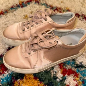 Ros färgade sneakers Axel Arigato satin - Snygga sneakers från Axel Arigato i en ljus beige/rosa ton med satinfinish. Skorna har rund tå, platt sula och snörning framtill. Perfekta för dig som gillar stilrena och trendiga detaljer. Märket syns diskret på sidan.