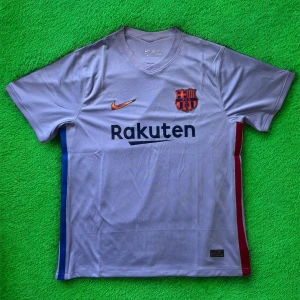 FC Barcelona bortatröja Nike M - Snygg FC Barcelona bortatröja från Nike i ljuslila polyester. Tröjan har korta ärmar, V-ringning och tryckta detaljer som klubbmärke, Nike-logga i kopparfärg och Rakuten-sponsor. Sidorna har blå och röda ränder, och Unicef-logga finns på ryggen.
