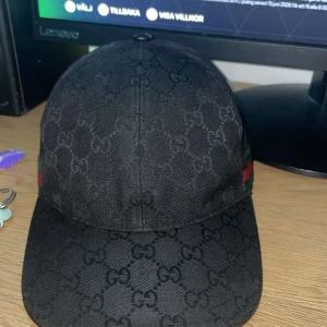 Svart keps från Gucci med GG-mönster - Snygg svart keps från Gucci med klassiskt GG-monogram över hela kepsen. På sidan finns en grön och röd rand som ger den en extra lyxig touch. Perfekt accessoar för att lyfta din outfit.
