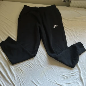 Svarta mjukisbyxor från Nike M - Svarta mjukisbyxor från Nike med klassisk vit logga på vänster ben. Byxorna har resår i midjan med snörning och muddar vid bensluten. Tillverkade i mjukt material som är perfekt för chill eller träning. Och har en bakficka som stäng med en knapp. Storlek M nypris 699
