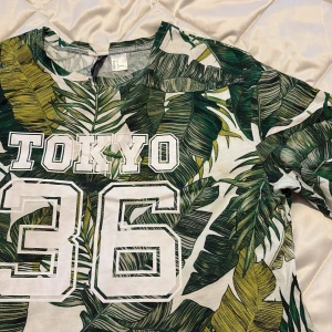 Grön Tokyo 36 klänning  från H&M - Snygg klänning  från Divided med stort tryck 'TOKYO 36' på bröstet. Klänningen har tropiskt bladmönster i grönt och gult på vit botten, rund hals och korta ärmar. 💚🌴