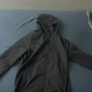 Svart vindjacka från Under Armour XS - Svart vindjacka från Under Armour i storlek XS. Jackan har huva, hel dragkedja framtill och diskret logga på bröstet samt stor logga på ryggen. Tillverkad i lätt och smidigt syntetmaterial, perfekt för aktiva dagar och träning. Den har en liten blå fläck,PRIS KAN DISKUTERAS 