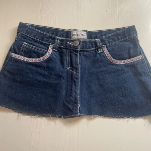 Blå jeanskjol med broderi från Check This - Supersöt kort jeanskjol i mörkblå denim från Check This. Kjolen har rå kant nedtill, broderade detaljer med blommor vid fickorna och coola bakfickor med vit söm.